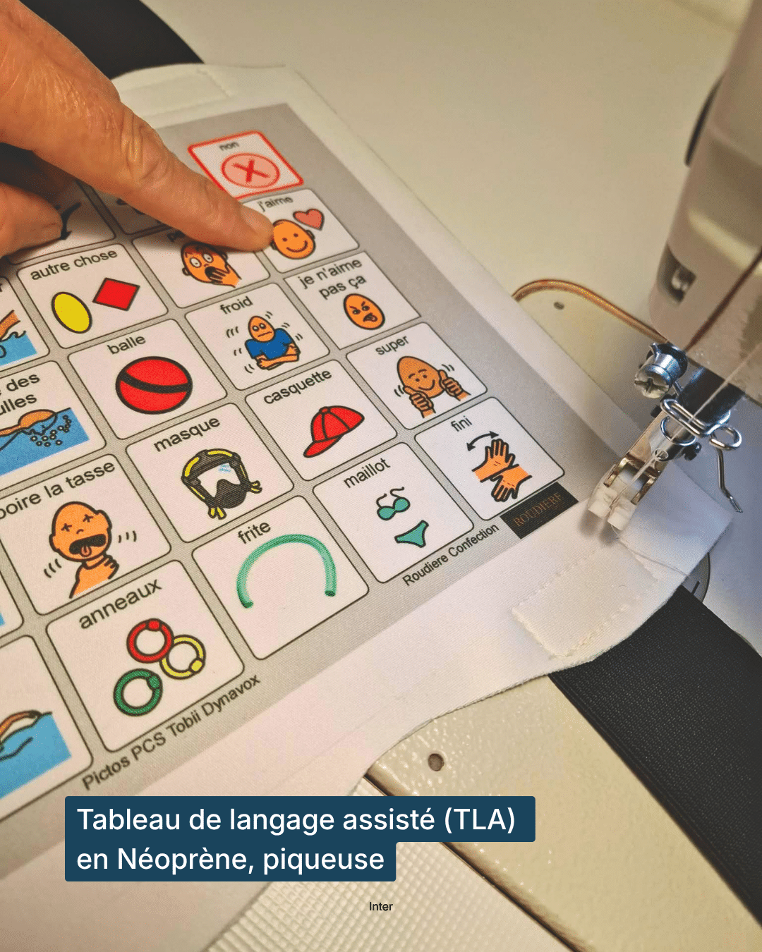 Gros plan sur un tableau de langage assisté en néoprène en cours de couture sur une machine à coudre, avec des pictogrammes colorés (oui, non, j’aime, je n’aime pas, froid, masque, casquette, maillot, balle, anneaux, fini) et un doigt qui en désigne un.