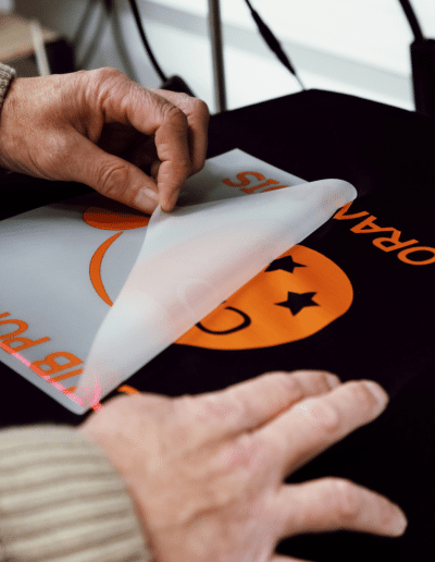 Mains qui retirent le film transparent d’un transfert DTF orange sur un textile noir.